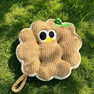 Braon Cuddle Puff jastuk u obliku kružića sa vezenim okicama, žutim kljunićem i zelenim listićem, na travi