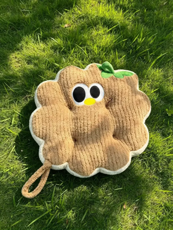 Braon Cuddle Puff jastuk u obliku kružića sa vezenim okicama, žutim kljunićem i zelenim listićem, na travi