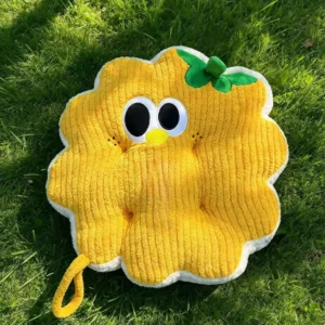 Žuti Cuddle Puff jastuk u obliku kružića sa vezenim okicama, kljunićem i zelenim listićem, na travi